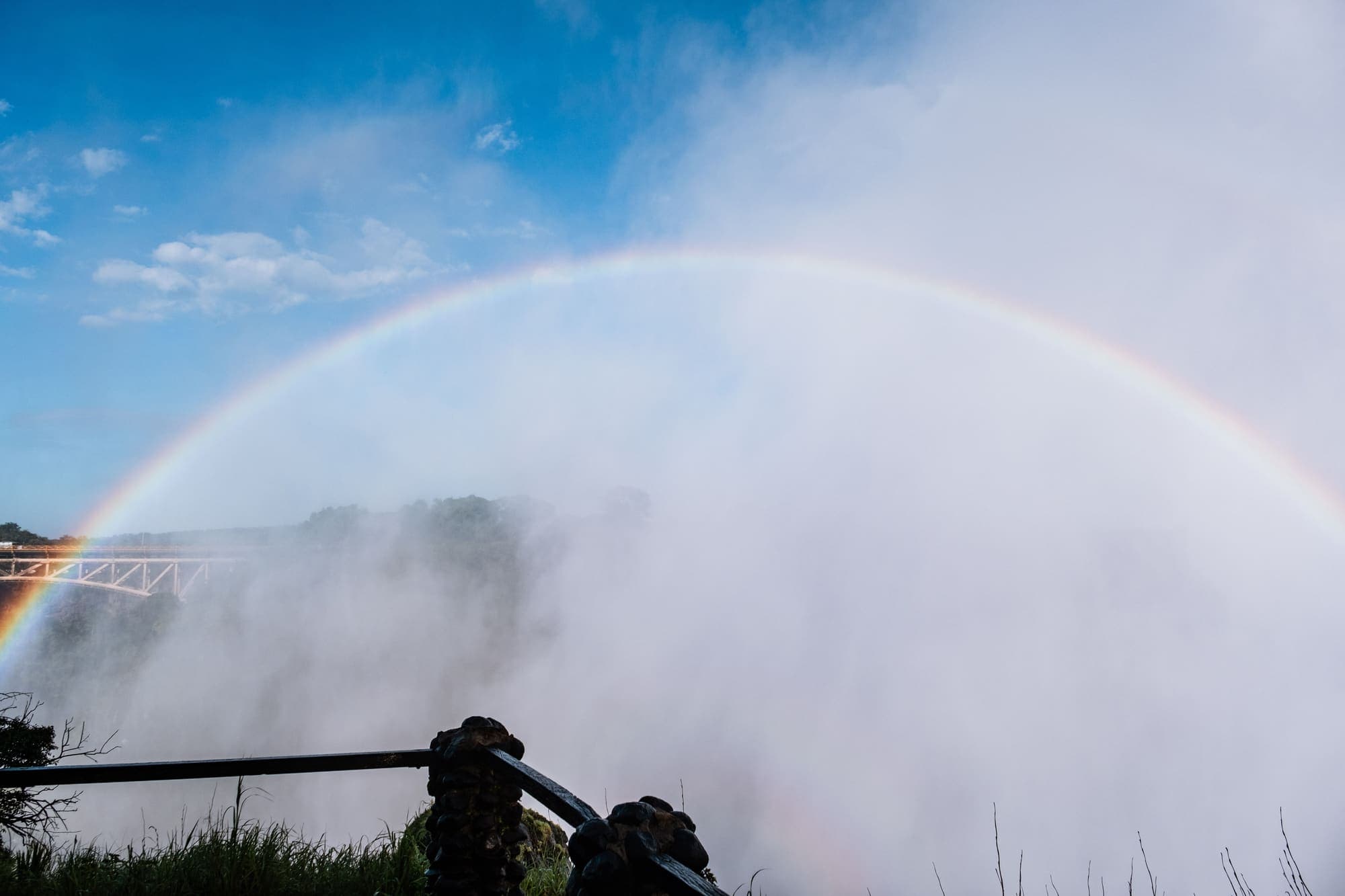 Arco-íris sobre a névoa das Cataratas Victoria, com a ponte entre o Zimbábue e a Zâmbia ao fundo, à esquerda.
