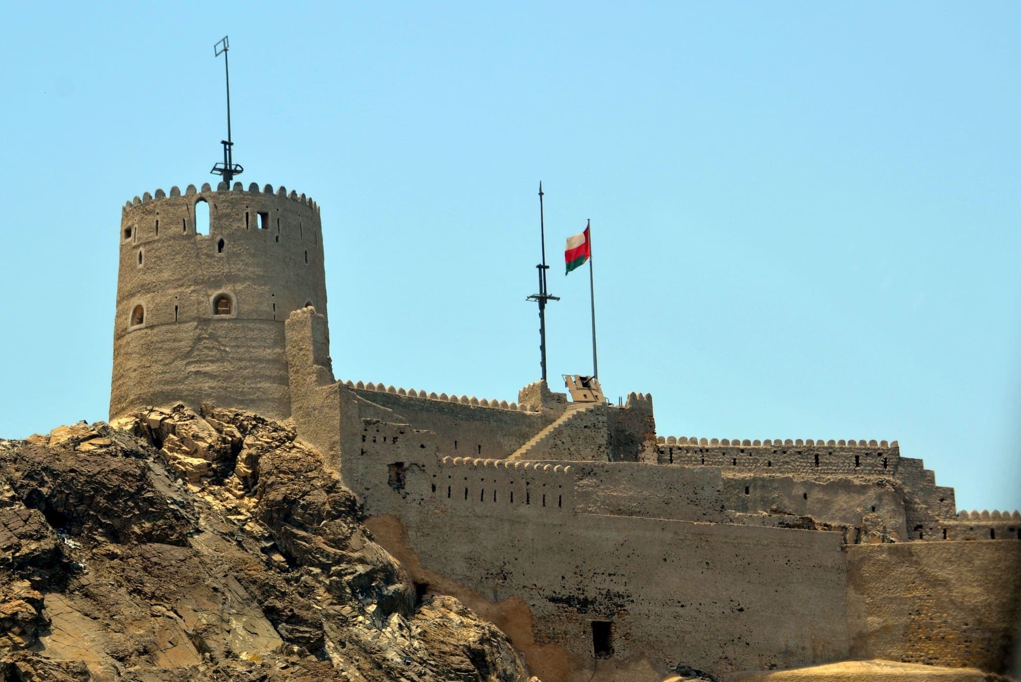 O Forte de Mutrah, em Mascate, com sua torre redonda e as muralhas sobre as rochas, e a bandeira de Omã ao lado.