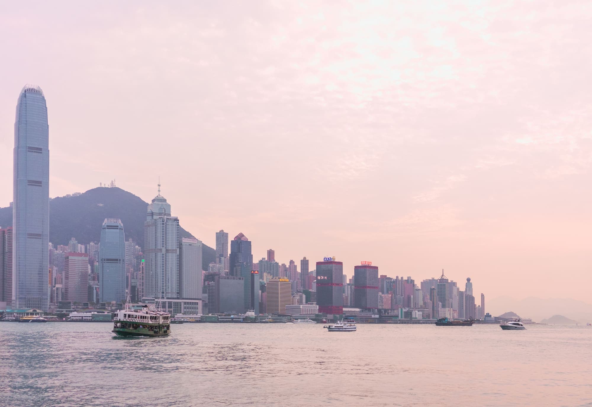 Os arranha-céus de Hong Kong à beira do porto Victoria, com uma balsa atravessando a baía ao entardecer.