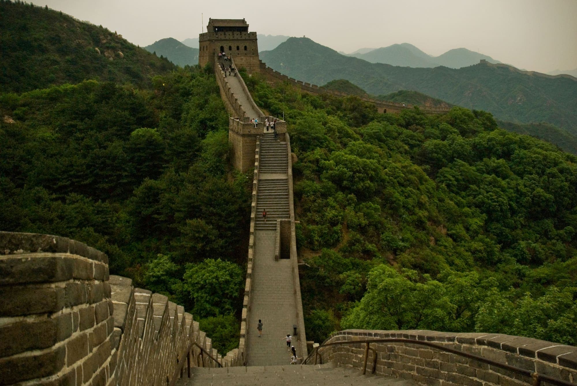 A Grande Muralha da China subindo um morro coberto de mata até uma torre de vigia.