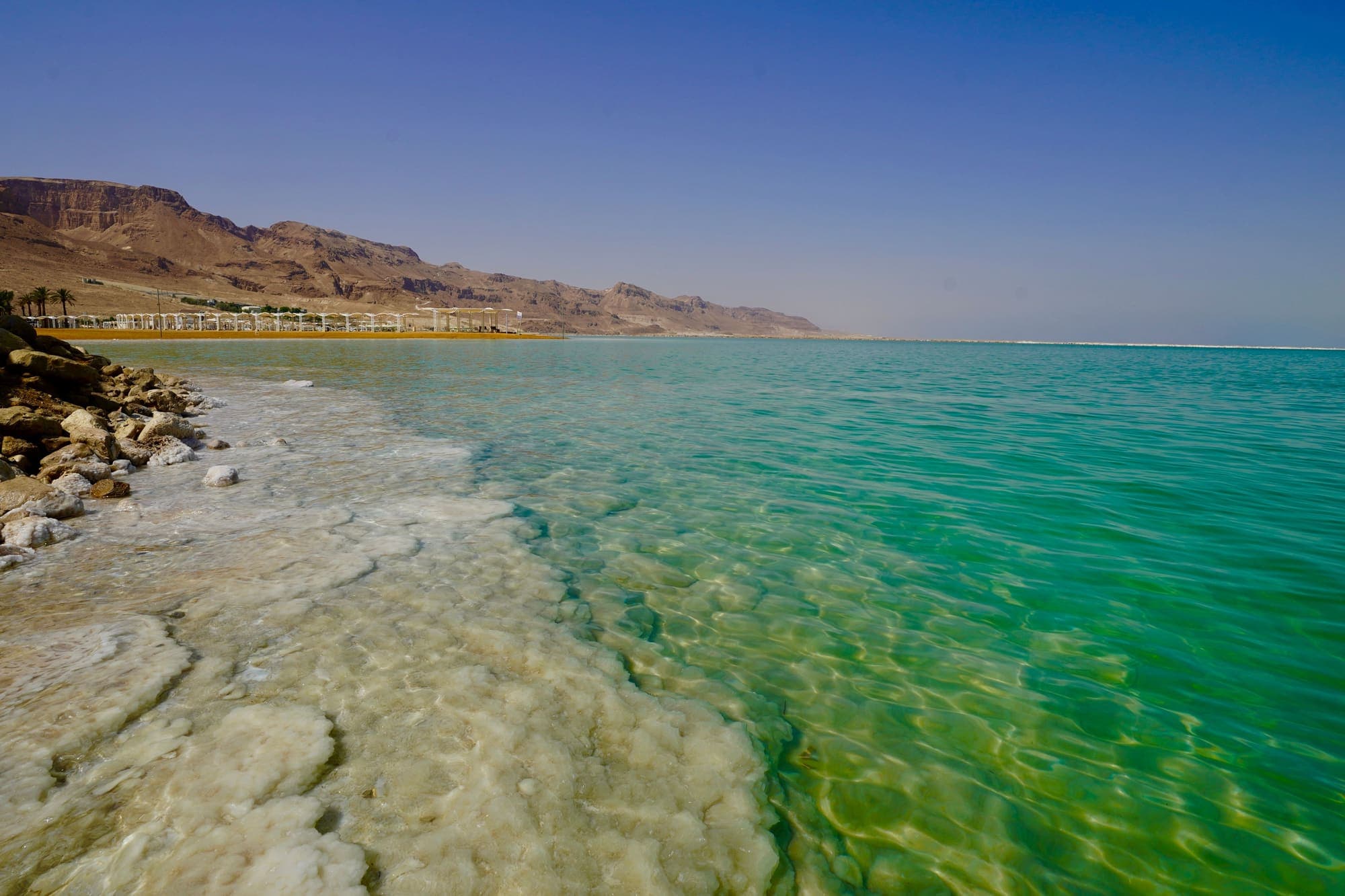 A margem do Mar Morto, com crostas de sal sob a água verde-turquesa e as montanhas do deserto ao fundo.