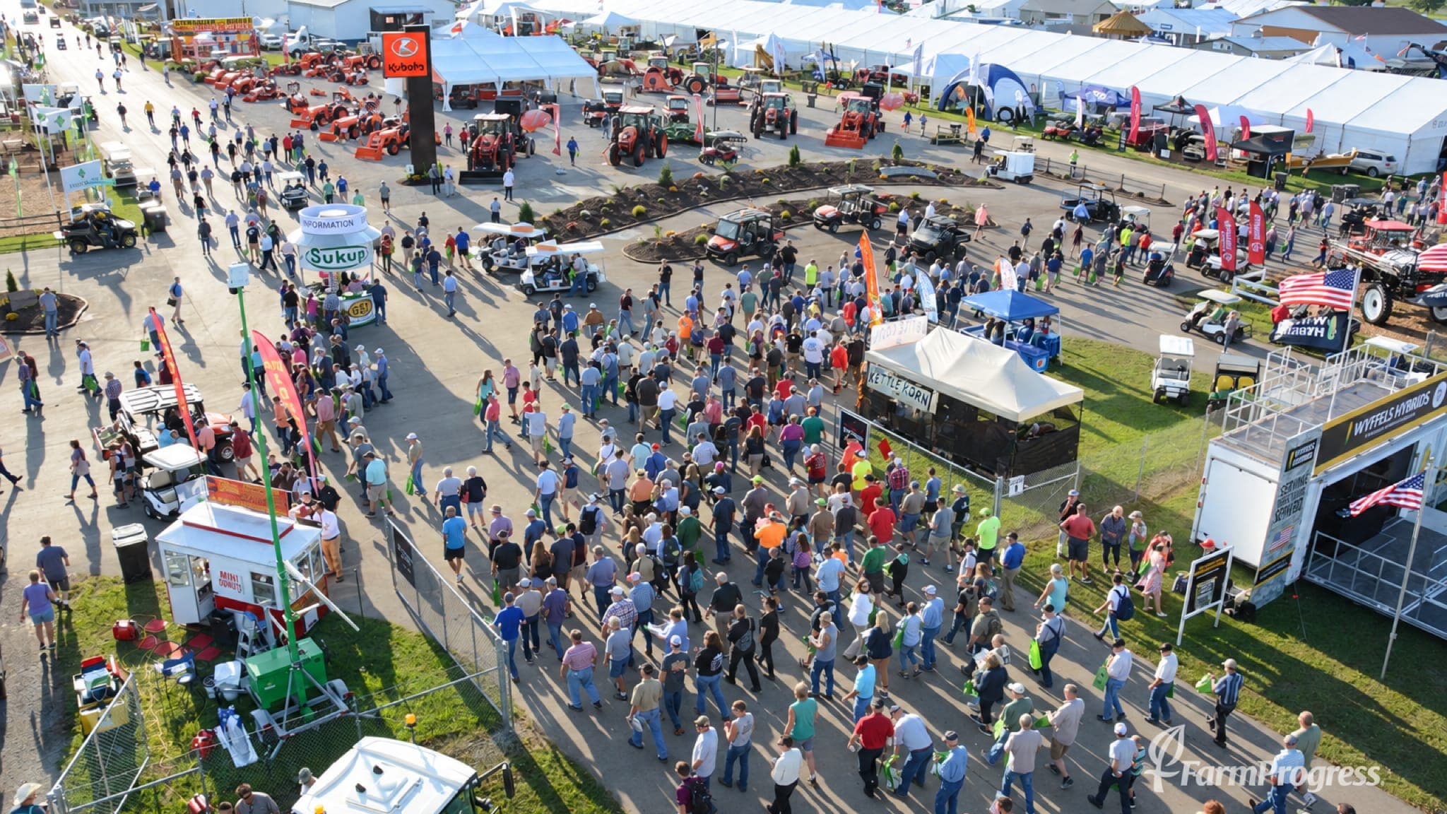 Vista do alto de visitantes caminhando entre os estandes, tratores e tendas da Farm Progress Show, num dia de sol.