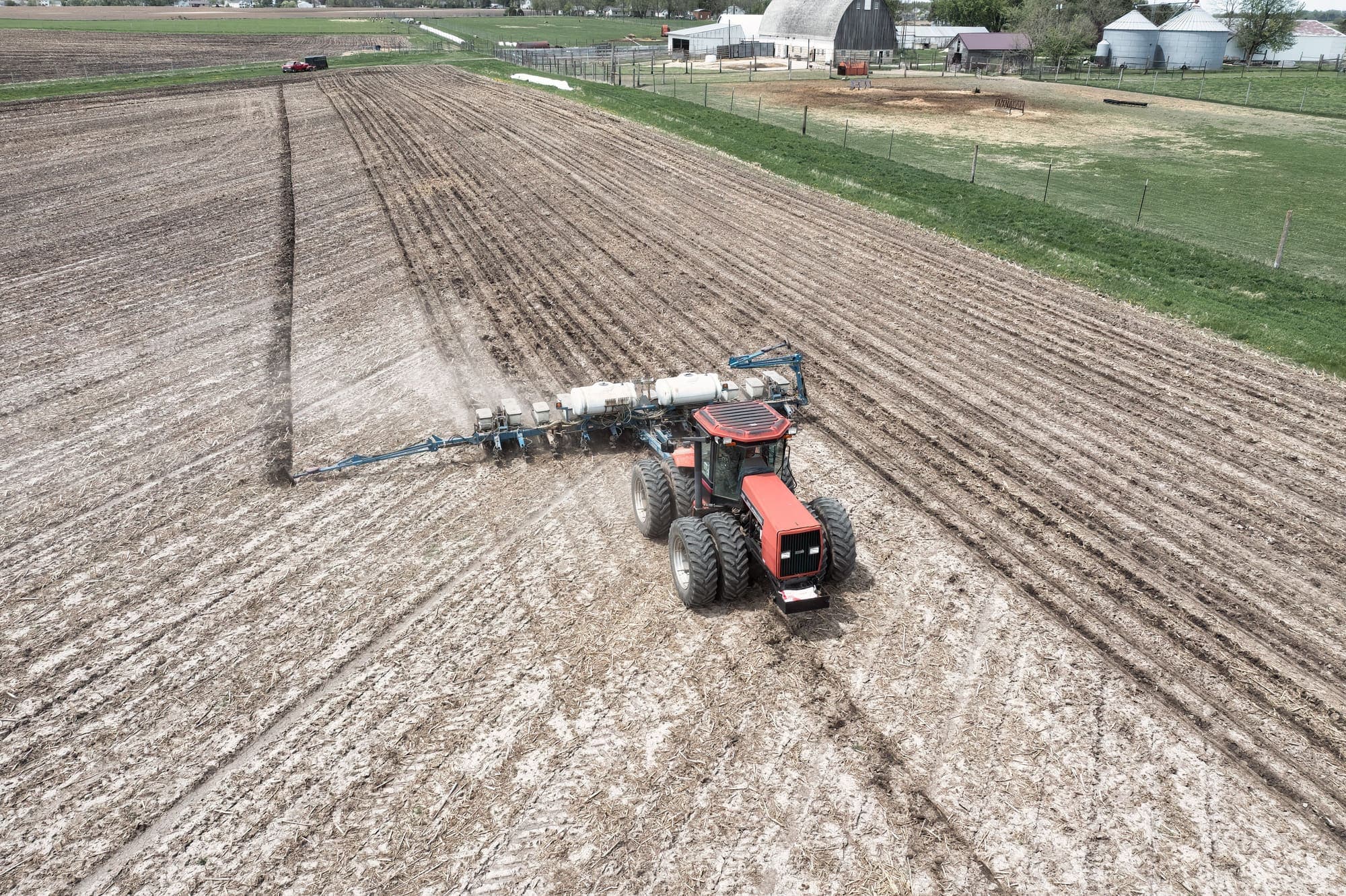 Trator puxando uma plantadeira de várias linhas num campo preparado, com os silos e galpões de uma fazenda ao fundo.