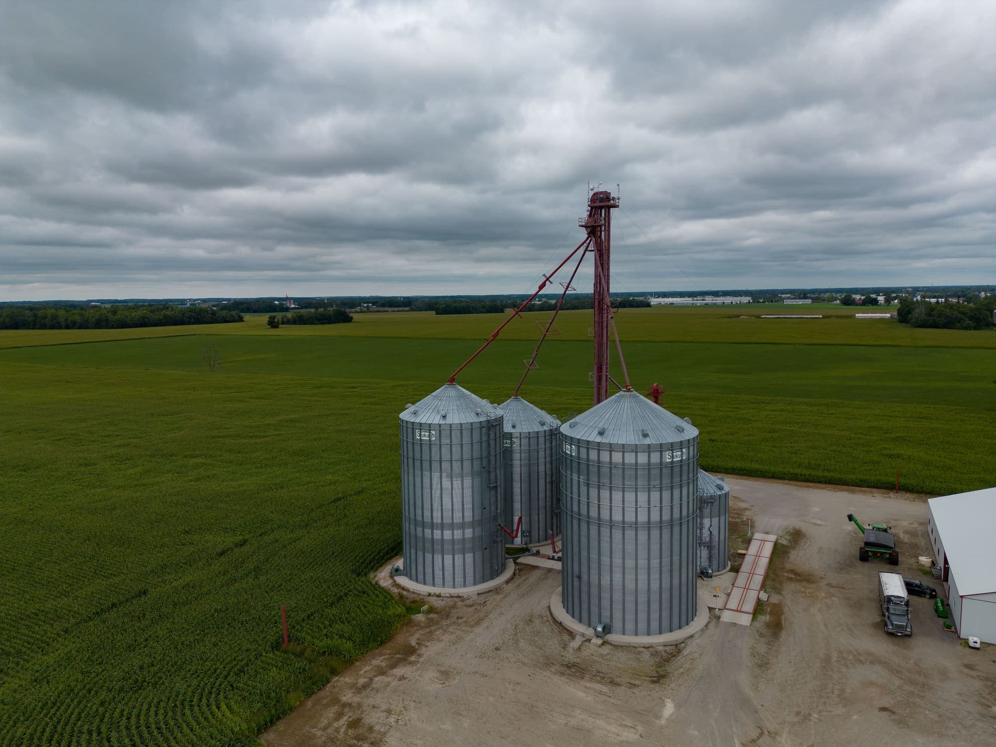Silos metálicos de grãos, com elevador e galpão de máquinas, numa fazenda cercada de lavouras verdes.
