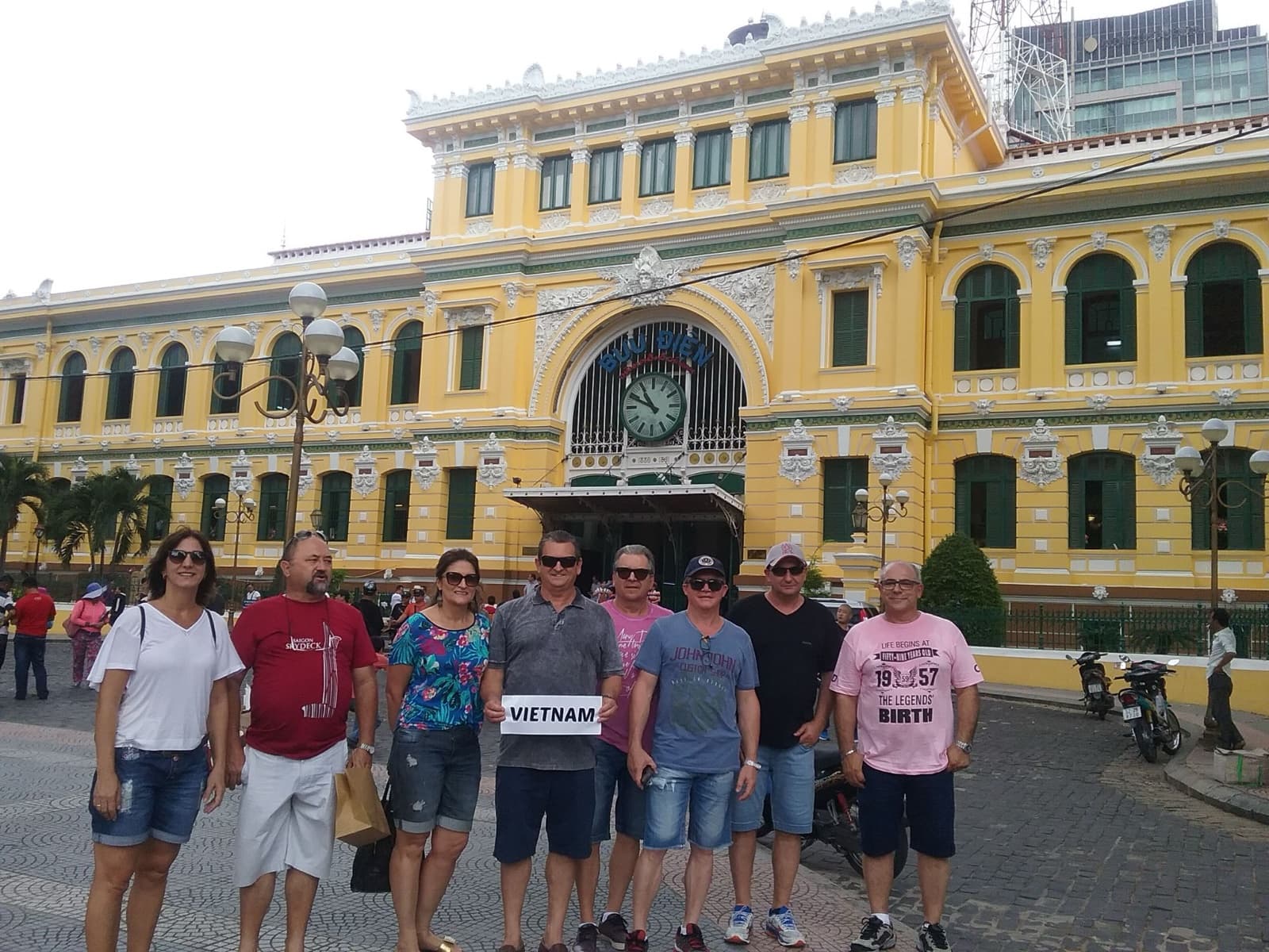 Grupo da Leão Viagens com um cartaz escrito Vietnam diante do prédio amarelo dos Correios de Saigon, no Vietnã.
