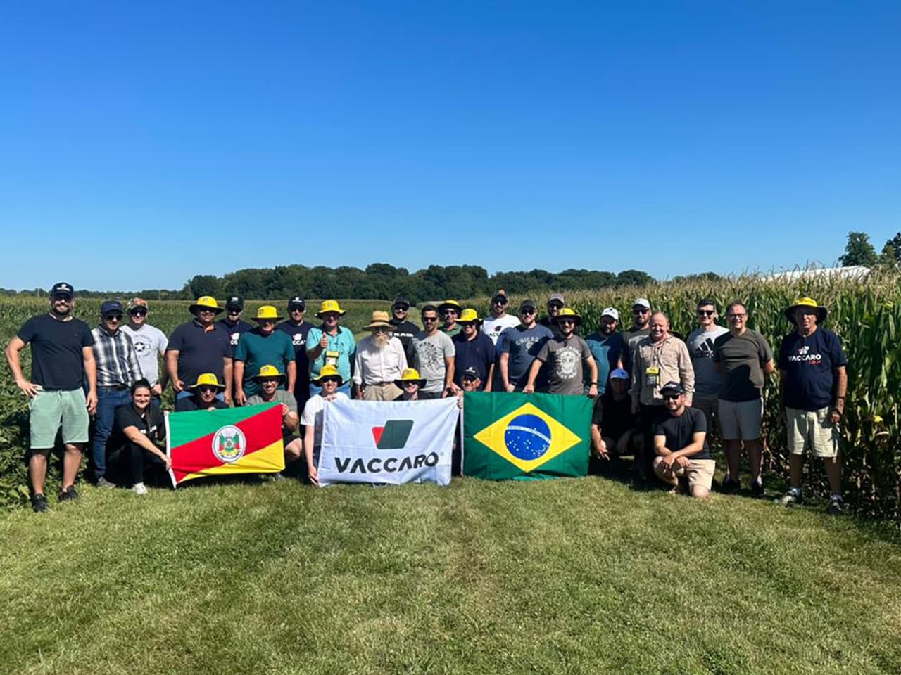 Grupo Vaccaro, muitos com o chapéu amarelo da Leão, diante de um milharal nos Estados Unidos, com as bandeiras do Rio Grande do Sul, da Vaccaro e do Brasil.